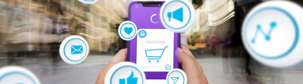 Two hands holding a smartphone in the middle of the road, displaying an online shopping cart and full of symbols for likes, ads and shares...