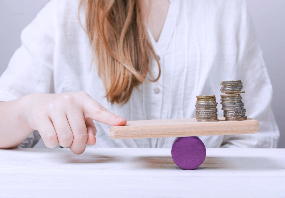 A woman sits in front of a table. A balance board is on a purple wheel. On one side of the board are coins, and on the other, the woman's hand.